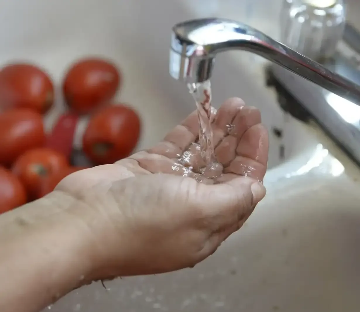 a person's hand positioned under a running tap, with clear water flowing over the fingers. The scene captures the simplicity and freshness of the water, emphasizing cleanliness and daily routine