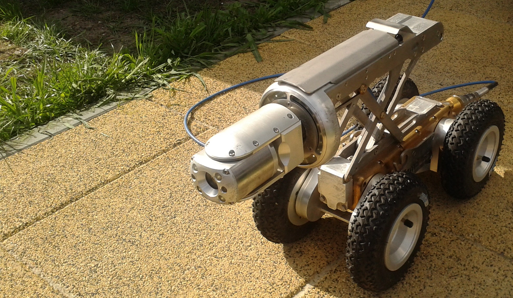 A stainless steel CCTV drain survey crawler with a camera head and four rubber tyres, connected to a blue cable, on a paved path beside a lawn
