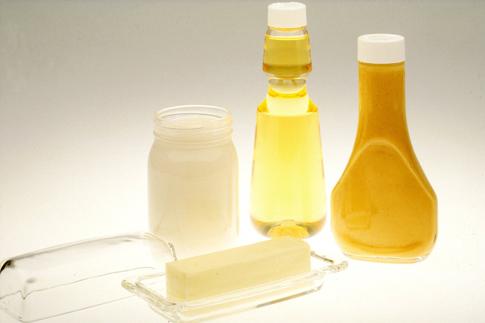 A glass jar of set white cooking fat beside bottles of vegetable oil and salad dressing and a block of butter